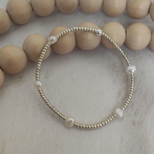以下が含まれることがあります： 5つの白い真珠が付いた、銀色のビーズブレスレット。 ブレスレットはゴムバンドに通されており、背景に木製のビーズがある白い表面に表示されています。