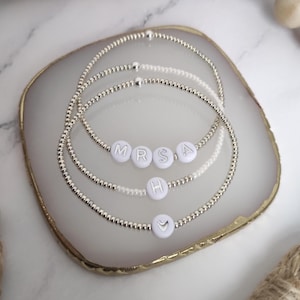 May include: Four beaded bracelets with white letter and symbol beads spelling "MRS A" and a heart. The bracelets are silver and sit on a white and gold-edged tray. The bracelets are a delicate and personalized accessory.