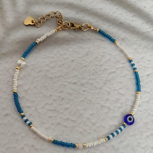 May include: Ankle bracelet with a beaded design in white, blue, and gold. It features a gold-coloured heart charm and an evil eye charm. The adjustable chain is gold-coloured.