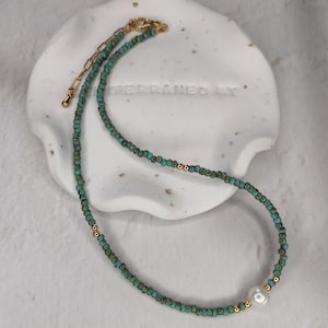 May include: A delicate turquoise beaded necklace with a single white pearl accent. The necklace is made with small, square-shaped beads and has a gold clasp.