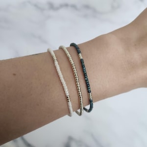 Puede incluir: Tres pulseras de cuentas en colores marfil, dorado y turquesa se muestran en una muñeca. Las pulseras están hechas de pequeñas cuentas ensartadas. La pulsera dorada está en el centro, flanqueada por las pulseras marfil y turquesa. Las pulseras son delicadas y minimalistas.