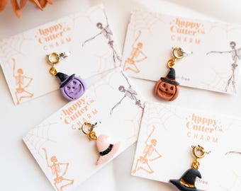 Könnte beinhalten: Halloween-Anhänger mit goldfarbenen Verschlüssen. Die Anhänger zeigen Miniatur-Hexenhüte, Kürbisse und eine lila Hexe. Jeder Anhänger ist an einer Karte mit dem Text "happy Cutter Co. CHARM" befestigt.