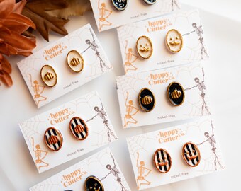 Könnte beinhalten: Eine Auswahl an Halloween-Ohrsteckern mit verschiedenen Designs, darunter Kürbisse, Skeletthände und Streifenmuster. Jedes Paar wird auf einer Karte mit dem Text "happy Cutter Co." und "nickel-free" präsentiert. Die Ohrringe sind oval.