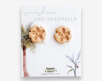 Könnte beinhalten: Ein Paar Sanddollar-Ohrringe in hellbrauner Farbe. Die Ohrringe werden auf einer weißen Karte mit dem Text "sunshine and seashells" und einem Logo für "happy cutter co." präsentiert.