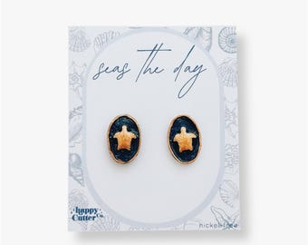 Könnte beinhalten: Ein Paar goldene und blaue Schildkröten-Ohrstecker auf einer weißen Karte mit dem Text "Seas the day" und "Happy Cutter Co." Die Karte trägt außerdem den Hinweis "nickel free".