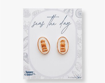 Könnte beinhalten: Ein Paar goldfarbene Leuchtturmohrringe auf einer weißen Karte mit dem Text "Seas the day" und "Happy Cutter Co." Die Karte trägt außerdem den Hinweis "nickel-free".