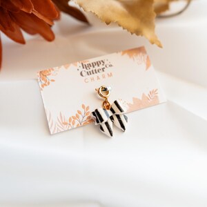 K&ouml;nnte beinhalten: Ein schwarz-wei&szlig; gestreifter Schleifenanh&auml;nger mit goldfarbenem Verschluss, befestigt an einer Karte mit der Aufschrift "happy Cutter Co. CHARM". Der Anh&auml;nger wird auf einem wei&szlig;en Stoffhintergrund mit herbstlichen Dekorationselementen pr&auml;sentiert.