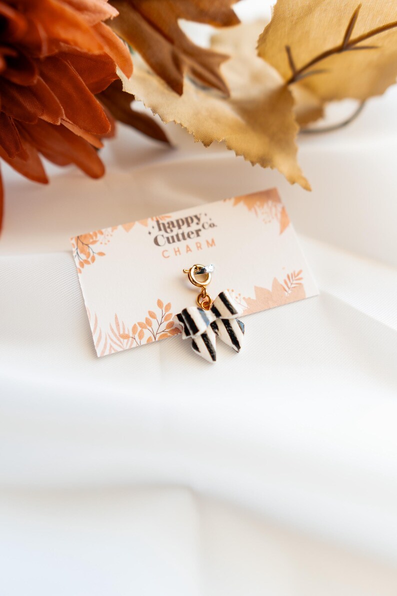 K&ouml;nnte beinhalten: Ein schwarz-wei&szlig; gestreifter Schleifenanh&auml;nger mit goldfarbenem Verschluss, befestigt an einer Karte mit der Aufschrift "happy Cutter Co. CHARM". Der Anh&auml;nger wird auf einem wei&szlig;en Stoffhintergrund mit herbstlichen Dekorationselementen pr&auml;sentiert.
