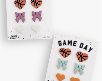 Könnte beinhalten: Zwei Ohrring-Sets auf weißen Karten. Auf jeder Karte steht oben der Text "GAME DAY". Eine Karte hat orangefarbene Basketball-Herz-Ohrringe, lila Schleifen-Ohrringe und weiße Herz-Ohrringe. Die andere Karte hat orangefarbene Basketball-Herz-Ohrringe, blaue Schleifen-Ohrringe und orangefarbene Herz-Ohrringe.