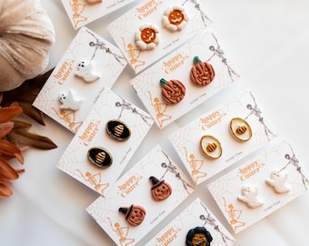 Könnte beinhalten: Eine Sammlung von Halloween-Ohrsteckern mit verschiedenen Designs, darunter Geister, Kürbisse und Hexenhüte. Die Ohrringe werden auf Karten mit dem Logo "happy cutter co." und dem Schriftzug "nickel-free" präsentiert. Die Ohrringe sind auf einer weißen Oberfläche angeordnet.