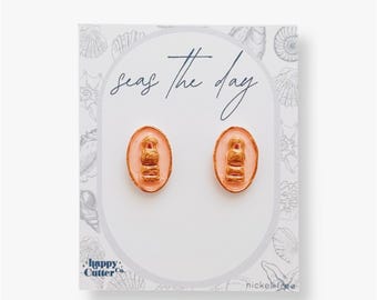 Könnte beinhalten: Ein Paar rosa und goldene Muschel-Ohrringe auf einer weißen Karte mit dem Text "Seas the day" und "Happy Cutter Co." Die Karte trägt außerdem den Hinweis "nickel free".