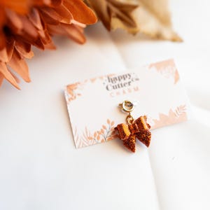 K&ouml;nnte beinhalten: Ein schleifenf&ouml;rmiger Charm mit goldfarbenem B&uuml;gel und glitzerndem orangefarbenem Finish, befestigt an einer Karte mit dem Text "happy cutter co. CHARM". Der Charm befindet sich vor einem wei&szlig;en Hintergrund mit orangefarbenen floralen Akzenten.