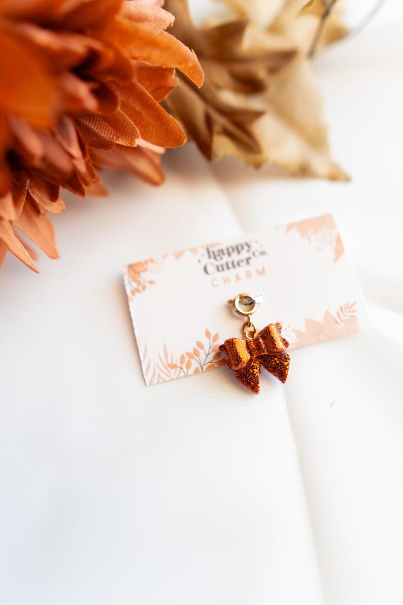 K&ouml;nnte beinhalten: Ein schleifenf&ouml;rmiger Charm mit goldfarbenem B&uuml;gel und glitzerndem orangefarbenem Finish, befestigt an einer Karte mit dem Text "happy cutter co. CHARM". Der Charm befindet sich vor einem wei&szlig;en Hintergrund mit orangefarbenen floralen Akzenten.