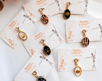 Könnte beinhalten: Ovale Halloween-Anhänger mit goldfarbenen Metallfassungen. Die Anhänger zeigen verschiedene Designs, darunter einen Kürbis, ein Jack-o'-Lantern-Gesicht und Streifenmuster. Jeder Anhänger ist an einer Karte mit dem Text "happy Cutter Co. CHARM" befestigt.