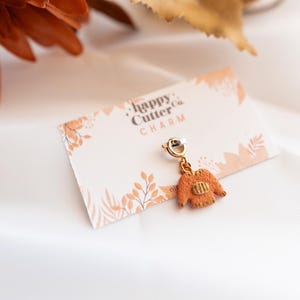 K&ouml;nnte beinhalten: Ein goldfarbener Anh&auml;nger in Form eines Miniatur-Orangepullovers, der an einem silbernen Verschluss h&auml;ngt. Der Anh&auml;nger befindet sich auf einer Karte mit dem Text "happy Cutter Co. CHARM" und einem floralen Design. Der Hintergrund ist ein wei&szlig;er, seidiger Stoff.