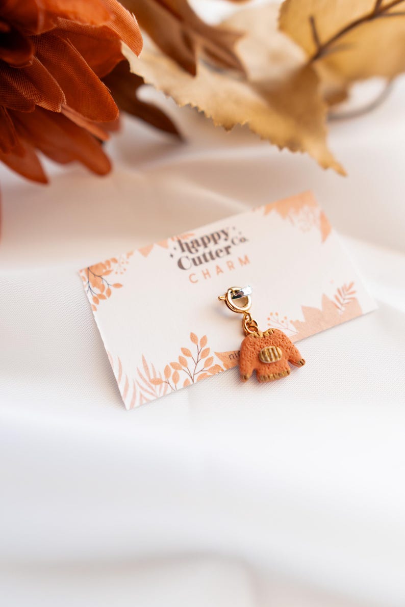 K&ouml;nnte beinhalten: Ein goldfarbener Anh&auml;nger in Form eines Miniatur-Orangepullovers, der an einem silbernen Verschluss h&auml;ngt. Der Anh&auml;nger befindet sich auf einer Karte mit dem Text "happy Cutter Co. CHARM" und einem floralen Design. Der Hintergrund ist ein wei&szlig;er, seidiger Stoff.