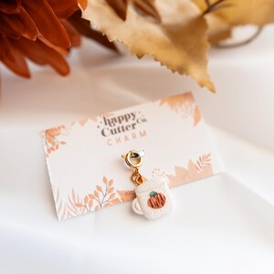 K&ouml;nnte beinhalten: Ein Charm mit einer kleinen wei&szlig;en Tasse mit einem K&uuml;rbismotiv, die an einem goldfarbenen Verschluss h&auml;ngt. Der Charm ist an einer Karte mit dem Text "happy Cutter co. CHARM" befestigt. Der Hintergrund umfasst herbstliche Dekorationen.