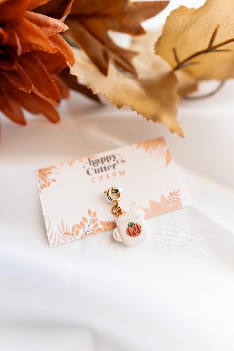 K&ouml;nnte beinhalten: Ein Charm mit einer kleinen wei&szlig;en Tasse mit einem K&uuml;rbismotiv, die an einem goldfarbenen Verschluss h&auml;ngt. Der Charm ist an einer Karte mit dem Text "happy Cutter co. CHARM" befestigt. Der Hintergrund umfasst herbstliche Dekorationen.