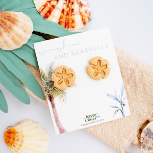 May include: A pair of gold-colored seashell earrings on a white card with the text "sunshine and seashells" and a palm tree illustration. The card also includes the text "Happy Cutter Co. happycutterco.com".