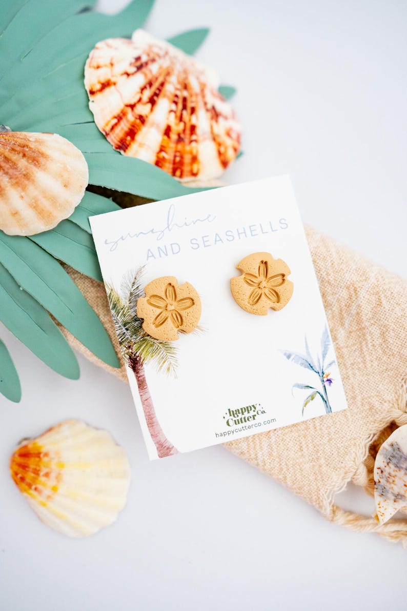 May include: A pair of gold-colored seashell earrings on a white card with the text "sunshine and seashells" and a palm tree illustration. The card also includes the text "Happy Cutter Co. happycutterco.com".
