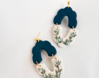 Könnte beinhalten: Ein Paar handgefertigte Ohrringe mit einem dunkelblauen, wolkenförmigen Oberteil und einem cremefarbenen, U-förmigen Unterteil. Der cremefarbene Bereich ist mit zarten, hellblauen floralen Akzenten verziert. Goldfarbene Ohrstecker vervollständigen das Design.