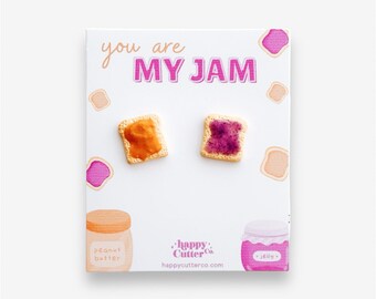 Könnte beinhalten: Eine weiße Karte mit dem Text "you are MY JAM" in rosa Buchstaben. Zwei kleine, realistisch aussehende Toaststücke mit Erdnussbutter und Marmelade befinden sich auf der Karte. Die Karte enthält auch ein kleines Glas Erdnussbutter und ein kleines Glas Marmelade.