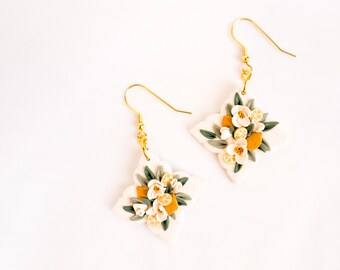 Könnte beinhalten: Ein Paar handgefertigte Ohrringe mit floralem Design. Jeder Ohrring hat eine weiße, vierzackige Form, die mit kleinen weißen Blüten, grünen Blättern und orangen Zitronenscheiben verziert ist. Die Ohrringe haben goldfarbene Haken.