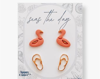 Könnte beinhalten: Ein Set mit vier Ohrringen mit einem pinkfarbenen Flamingo- und einem goldenen Flip-Flop-Design. Die Ohrringe werden auf einer weißen Karte mit dem Text "Seas the day" und "Happy Cutter Co." präsentiert.