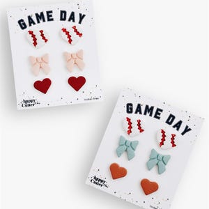Könnte beinhalten: Zwei Karten mit dekorativen Verzierungen. Jede Karte trägt den Text "GAME DAY" in Marineblau. Die Verzierungen umfassen Basebälle, Schleifen und Herzen in verschiedenen Farben. Der Markenname "happy cutter" ist sichtbar.
