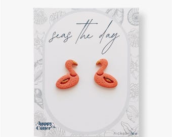 Könnte beinhalten: Ein Paar rosa Flamingo-Ohrringe mit goldenen Akzenten auf einer weißen Karte mit dem Text "Seas the day" und "Happy Cutter Co." in blauer Schrift. Die Karte trägt außerdem den Hinweis "nickel-free".