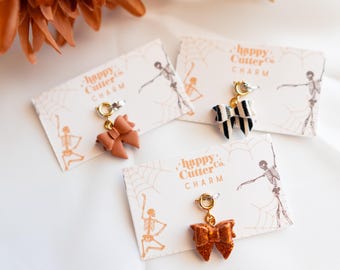 Könnte beinhalten: Drei schleifenförmige Anhänger in Braun-, Schwarz-Weiß- und Orangetönen, jeweils an einem goldfarbenen Verschluss befestigt. Die Anhänger werden auf Karten mit dem Text "happy Cutter Co. Charm" und einer Skelettgrafik präsentiert.
