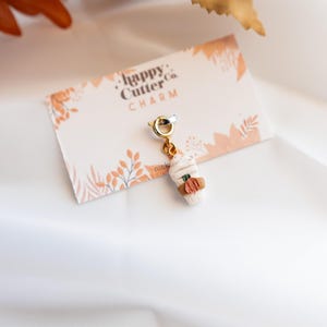 K&ouml;nnte beinhalten: Ein Miniatur-Charm in Form eines Kaffeebechers mit wei&szlig;em Schlagsahne-Topping und brauner H&uuml;lle. Der Charm hat ein kleines orangefarbenes K&uuml;rbisdetail und ist an einem goldfarbenen Verschluss befestigt. Die Karte tr&auml;gt die Aufschrift "happy Cutter Co. CHARM".
