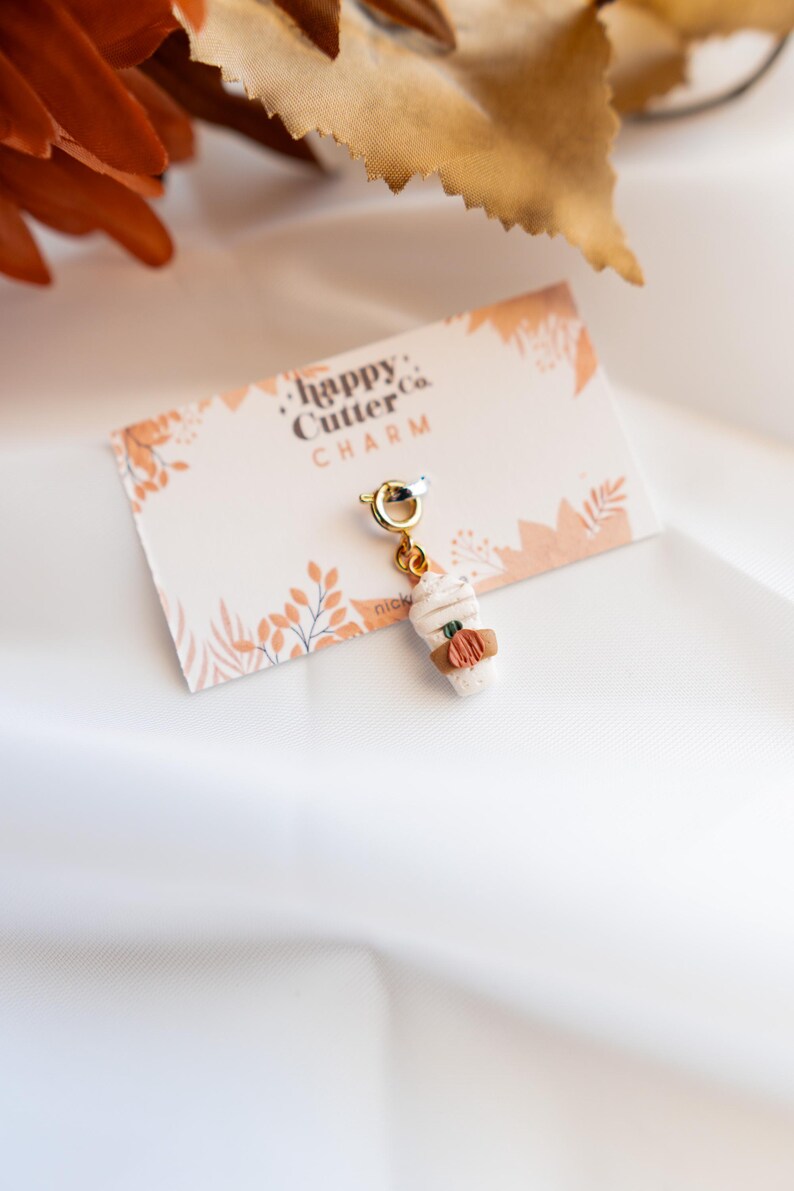 K&ouml;nnte beinhalten: Ein Miniatur-Charm in Form eines Kaffeebechers mit wei&szlig;em Schlagsahne-Topping und brauner H&uuml;lle. Der Charm hat ein kleines orangefarbenes K&uuml;rbisdetail und ist an einem goldfarbenen Verschluss befestigt. Die Karte tr&auml;gt die Aufschrift "happy Cutter Co. CHARM".