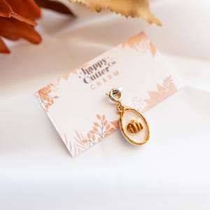 K&ouml;nnte beinhalten: Ein goldfarbener Charm mit einem K&uuml;rbismotiv auf wei&szlig;em Hintergrund. Der Charm ist an einer kleinen Karte mit dem Text "happy Cutter co. CHARM" und einem floralen Design befestigt. Der Hintergrund ist wei&szlig;.