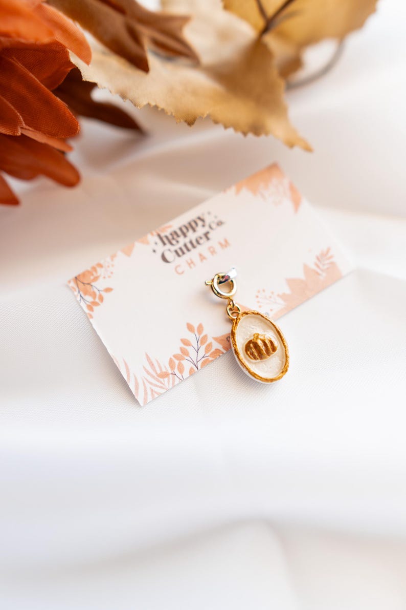K&ouml;nnte beinhalten: Ein goldfarbener Charm mit einem K&uuml;rbismotiv auf wei&szlig;em Hintergrund. Der Charm ist an einer kleinen Karte mit dem Text "happy Cutter co. CHARM" und einem floralen Design befestigt. Der Hintergrund ist wei&szlig;.