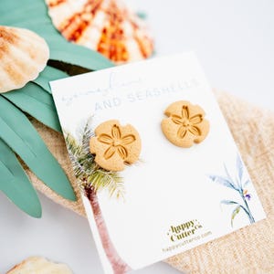 May include: A pair of sand dollar earrings in a light brown color. The earrings are displayed on a white card with the text "sunshine and seashells" and the logo "happy cutter co."