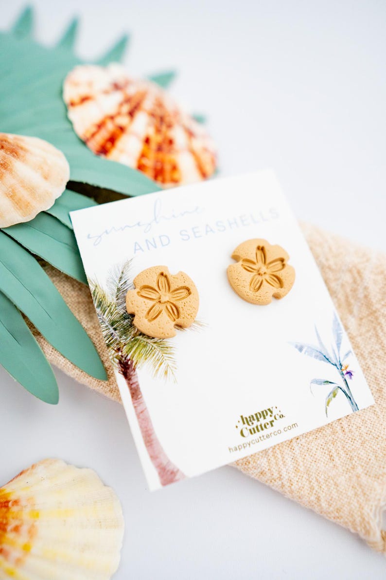 May include: A pair of sand dollar earrings in a light brown color. The earrings are displayed on a white card with the text "sunshine and seashells" and the logo "happy cutter co."