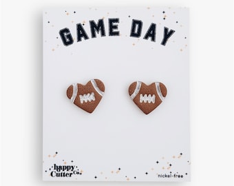 Könnte beinhalten: Herzförmige Ohrringe im Design von American Footballs. Die Ohrringe sind braun mit weißen Details, vor einem weißen Hintergrund mit dem Schriftzug "GAME DAY" in Marineblau. Der Markenname "happy cutter co." ist ebenfalls sichtbar.