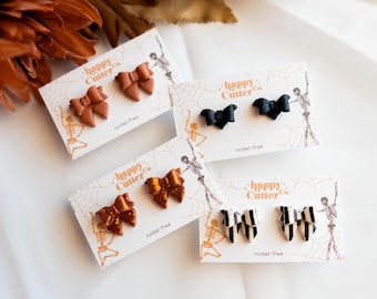 Könnte beinhalten: Vier Paar Ohrstecker auf Karten. Die Ohrringe sind wie Schleifen, Fledermäuse und gestreifte Schleifen geformt. Die Schleifen sind braun, orangefarben glitzernd, schwarz und schwarz-weiß. Auf den Karten steht "happy Cutter Co." und "nickel-free."