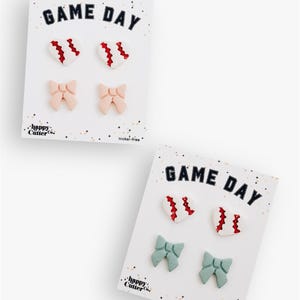 Könnte beinhalten: Zwei Karten mit Ohrringen. Jede Karte trägt den Text "GAME DAY" in Marineblau und Weiß. Eine Karte hat zwei weiße Baseball-Herz-Ohrringe mit roten Nähten und zwei rosa Schleifenohrringe. Die andere Karte hat die gleichen Baseball-Herz-Ohrringe und zwei türkisfarbene Schleifenohrringe.