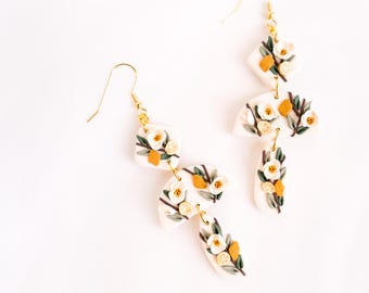 Könnte beinhalten: Handgefertigte Blumenohrringe in Cremefarbe mit goldfarbenen Haken. Die Ohrringe haben weiße Blüten, gelbe Akzente und grüne Blätter. Das Design ist ein dreidimensionales Blumenarrangement.