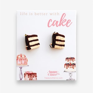 May include: A pair of polymer clay earrings shaped like a slice of chocolate cake with white frosting and sprinkles. The earrings are on a white card with the text "life is better with cake" and a logo for "happy cutter co."