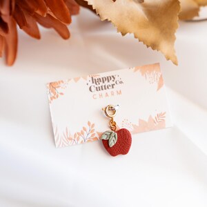 K&ouml;nnte beinhalten: Ein roter Apfel-Anh&auml;nger mit gr&uuml;nen Bl&auml;ttern h&auml;ngt an einem goldfarbenen Verschluss. Der Anh&auml;nger befindet sich auf einer Karte mit der Aufschrift "happy Cutter Co. CHARM". Der Hintergrund umfasst Herbstbl&auml;tter und eine wei&szlig;e Oberfl&auml;che.