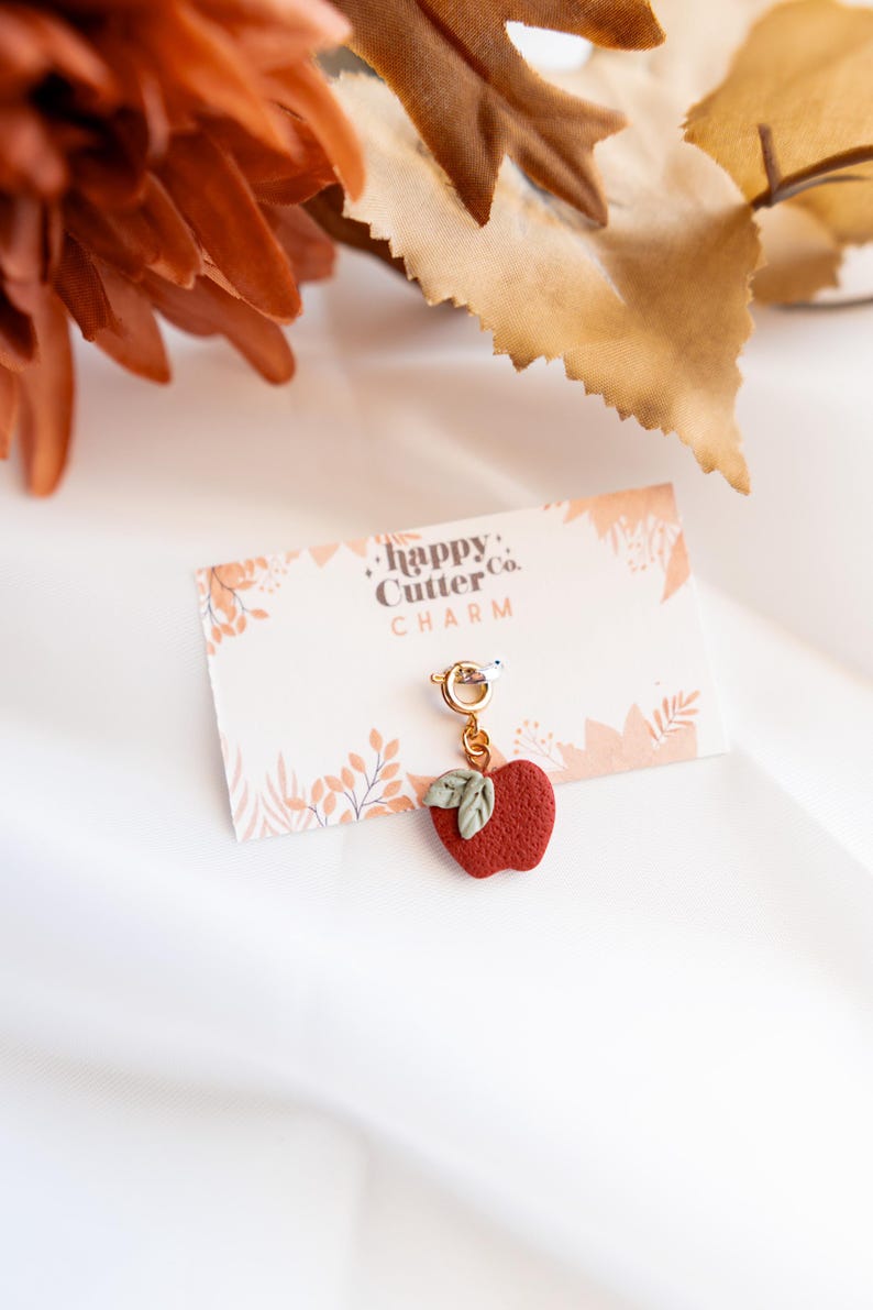K&ouml;nnte beinhalten: Ein roter Apfel-Anh&auml;nger mit gr&uuml;nen Bl&auml;ttern h&auml;ngt an einem goldfarbenen Verschluss. Der Anh&auml;nger befindet sich auf einer Karte mit der Aufschrift "happy Cutter Co. CHARM". Der Hintergrund umfasst Herbstbl&auml;tter und eine wei&szlig;e Oberfl&auml;che.