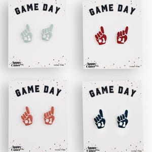 Könnte beinhalten: Vier Paar "Game Day" Ohrringe auf weißen Karten. Jedes Paar zeigt ein Schaumstofffinger-Design in verschiedenen Farben: Hellblau, Rot, Koralle und Marineblau. Die Karten zeigen auch den Text "happy Cutter Co." und "nickel-free."
