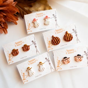 Könnte beinhalten: Eine Auswahl an Halloween-Ohrringen. Die Ohrringe zeigen Geister, Kürbisse und Jack-o'-Laternen, jeweils mit Hut. Die Ohrringe sind auf Karten mit dem Text "happy Cutter Co." und "nickel-free" präsentiert.