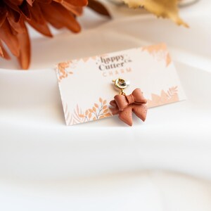 K&ouml;nnte beinhalten: Ein terrakottafarbener Schleifenanh&auml;nger mit einem goldfarbenen Verschluss, befestigt an einer Karte mit der Aufschrift "happy cutter CHARM". Der Anh&auml;nger befindet sich vor einem wei&szlig;en Hintergrund mit herbstlichen Dekorationselementen.