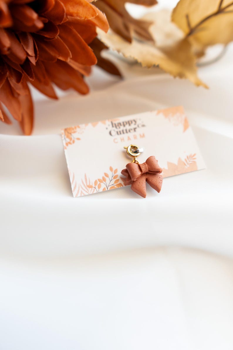 K&ouml;nnte beinhalten: Ein terrakottafarbener Schleifenanh&auml;nger mit einem goldfarbenen Verschluss, befestigt an einer Karte mit der Aufschrift "happy cutter CHARM". Der Anh&auml;nger befindet sich vor einem wei&szlig;en Hintergrund mit herbstlichen Dekorationselementen.