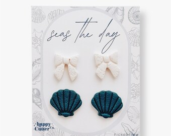Könnte beinhalten: Ein Set mit vier Ohrringen, bestehend aus zwei weißen Schleifen und zwei türkisfarbenen Muscheln. Die Ohrringe werden auf einer weißen Karte mit dem Text "Seas the Day" und "Happy Cutter Co." präsentiert. Die Karte trägt außerdem den Hinweis "nickel-free".