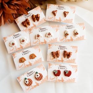 Peut inclure: Collection de boucles d'oreilles faites à la main en pâte polymère. Les boucles d'oreilles présentent divers motifs sur le thème de l'automne, notamment des nœuds, des citrouilles, des pulls et des pommes. Chaque paire est présentée sur une carte avec le texte "happy Cutter Co."